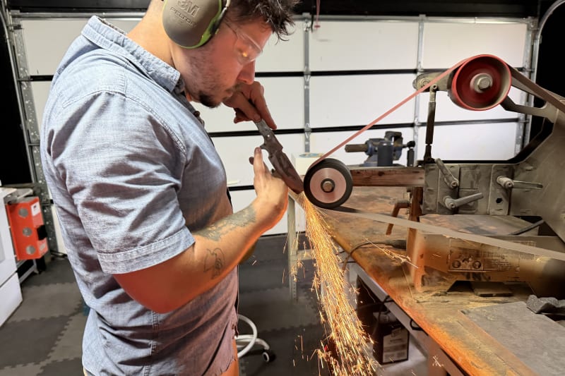 A close-up view of Dean Shroka, Severed Metal Works, in a workshop wearing safety glasses and ear protection, intently grinding a knife blade on a large belt grinder as sparks fly.
