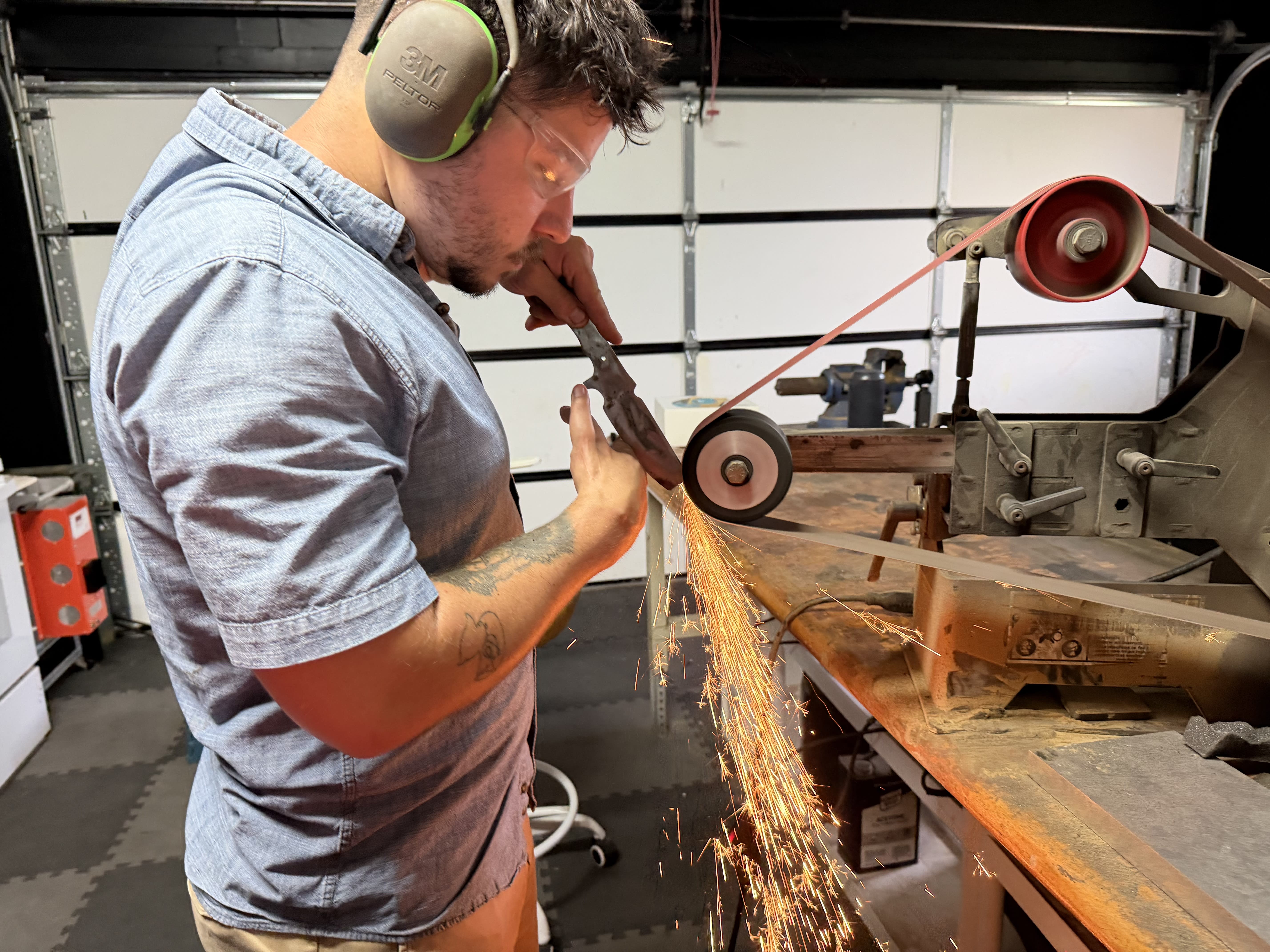 A close-up view of Dean Shroka, Severed Metal Works, in a workshop wearing safety glasses and ear protection, intently grinding a knife blade on a large belt grinder as sparks fly.