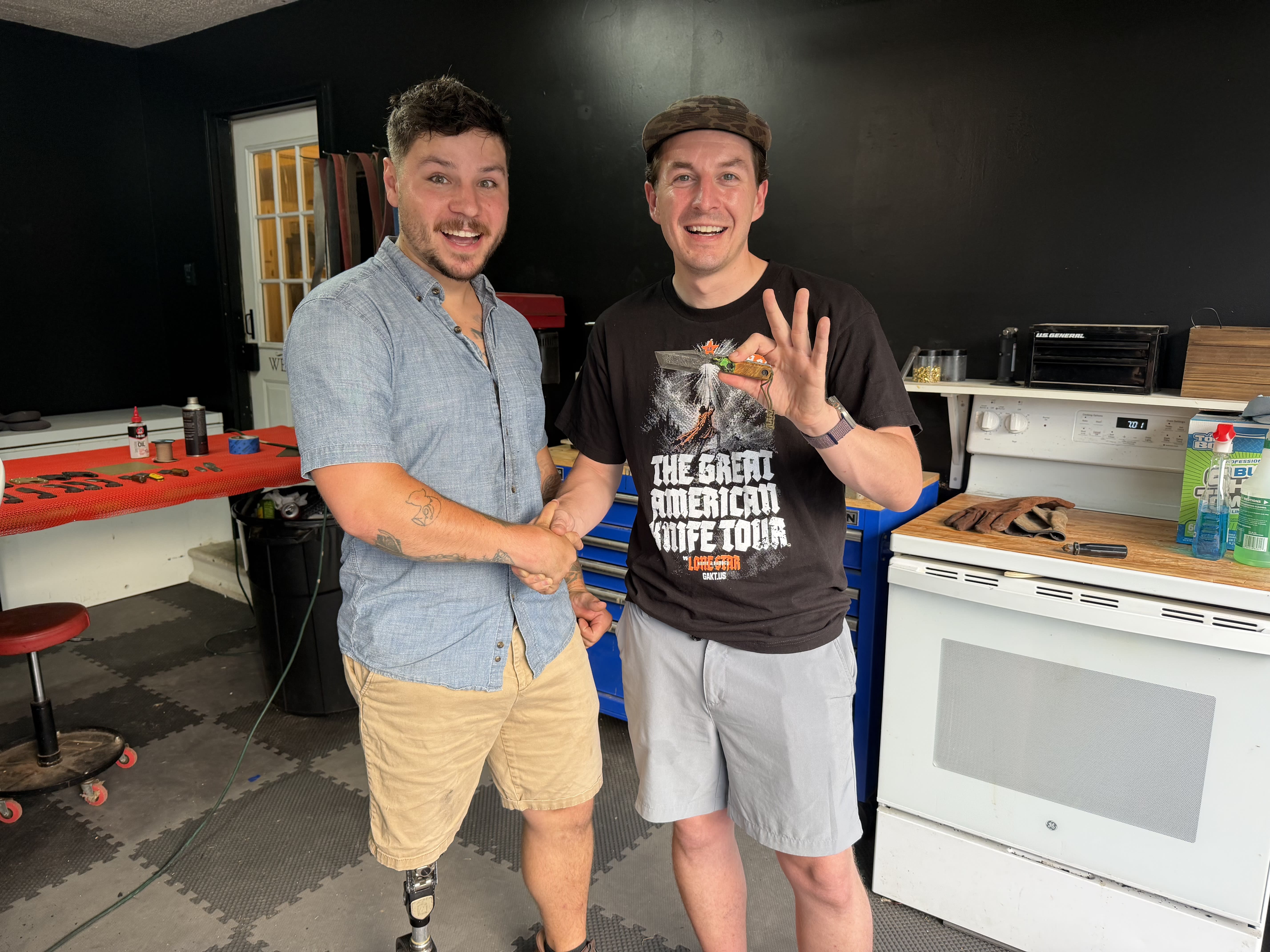 Two smiling men shaking hands inside a workshop; the man on the left wears a light blue button-down shirt and khaki shorts, and the man on the right wears a black graphic t-shirt and grey shorts, proudly holding up a custom knife.