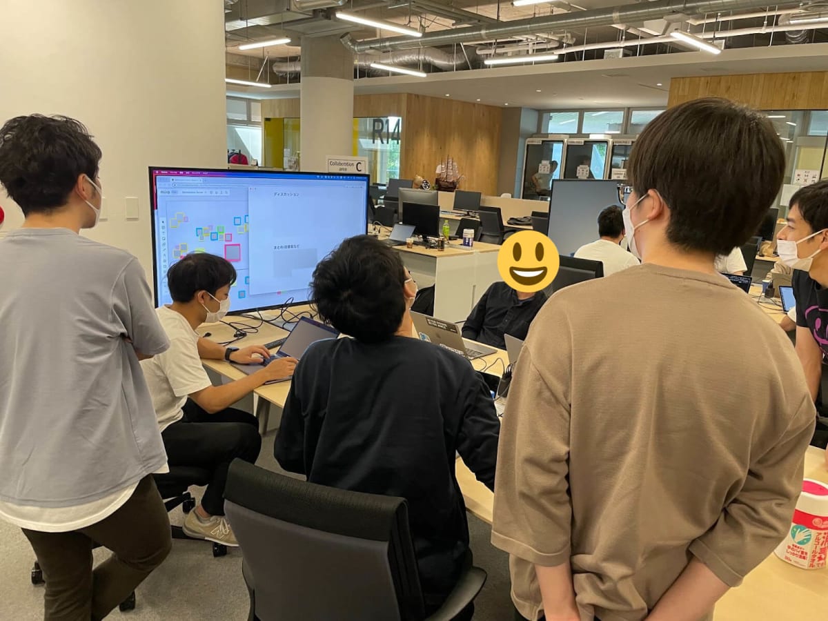 ハコベル株式会社のオフィス内で、自社プロダクトを触りながら複数のチームのエンジニアメンバーが一つになって打ち合わせをしている様子。ドキュメントツールを画面に投影し、気がついたことや課題などを書き込んでいる