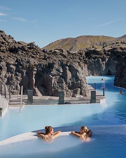 Two women relax in geothermal lagoon surrounded by rocky cliffs.