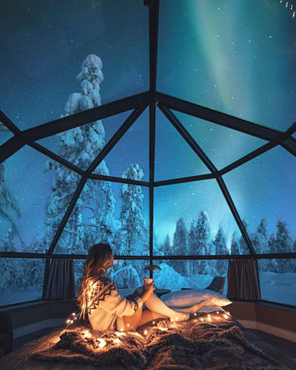 Woman in igloo cabin watches snowy forest and Northern Lights.