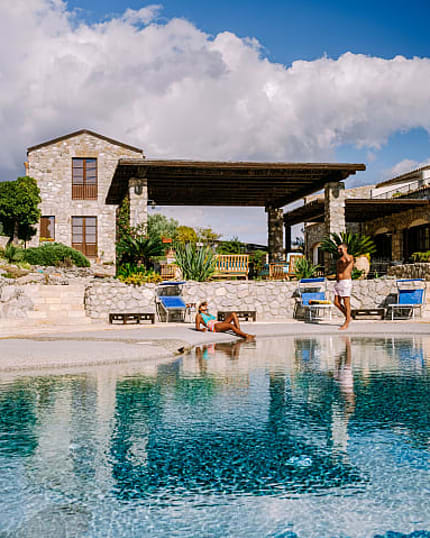 Couple relaxes by stone villa pool under bright blue sky.