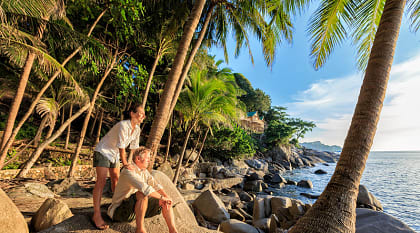 Senior couple enjoying a tropical beach at sunset surrounded by palm trees and rocky shoreline in Thailand.