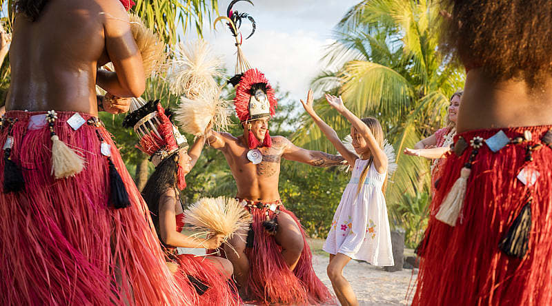 Photo courtesy of Tahiti Tourisme Grégoire Le Bacon Young girl and her mother with Tahitian men and women in traditional attire performing a cultural dance surrounded by lush greenery and a serene ocean backdrop in Tahiti