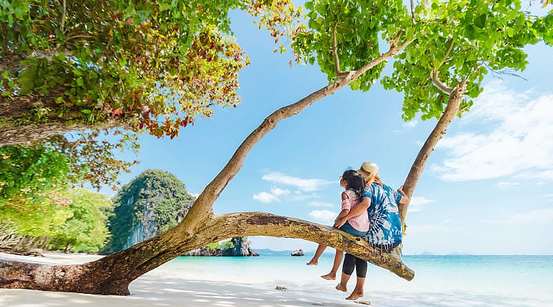 Mother and daughter sitting on a low tree branch on a beach in Phuket, Thailand