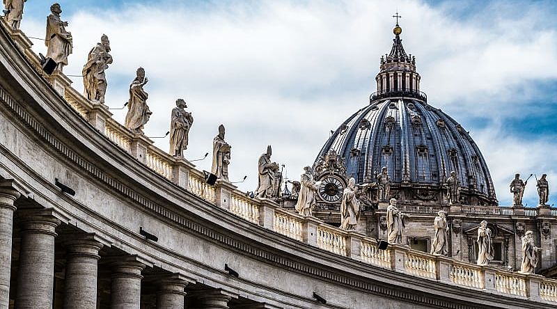 Intricate statues standing on a rooftop with the peak of St Peter's Basilica in the background.