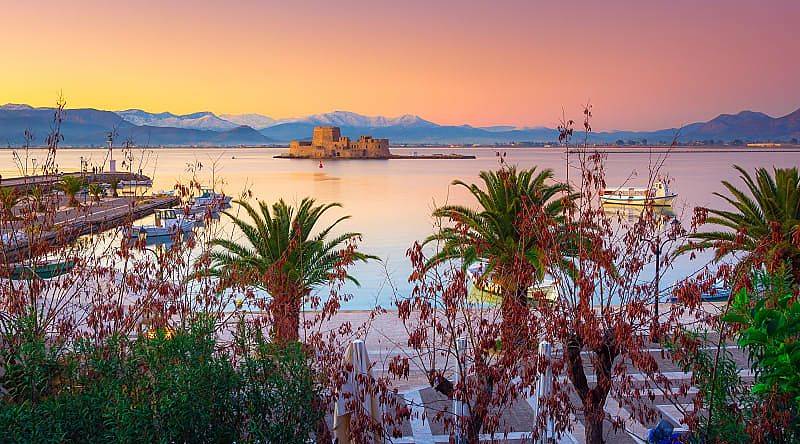 Nafplio Greece with palm trees, boats and Bourtzi castle on the water at sunset