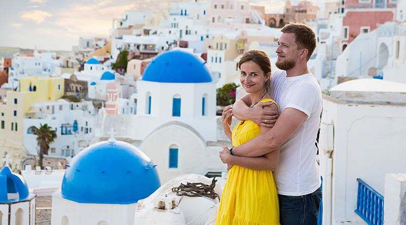 A couple enjoy the view amongst the white and pastel buildings, with famous blue domes in Santorini, Greece