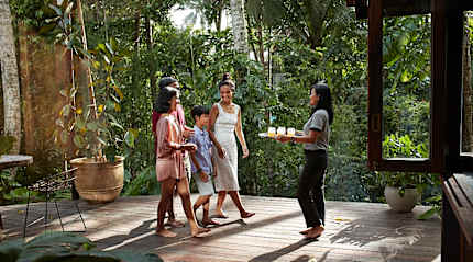 A female specialist stands on a sunny tropical balcony, smiling and gesturing while speaking to a traveling family.