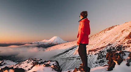 Hiker in winter jacket standing on snowy mountain, admiring view of snow-capped peaks and clouds at sunrise in Tongariro National Park, New Zealand
