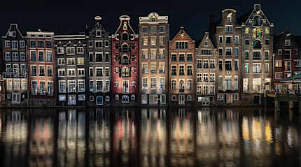 Illuminated traditional Dutch gabled houses reflecting in an Amsterdam canal at night.