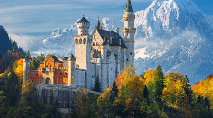 Neuschwanstein castle surrounded by trees with autumn colors and snowcapped mountain