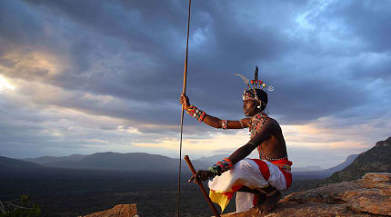 A traditional warrior sitting on a rocky hill at sunset, holding a spear and overlooking a vast wilderness landscape.