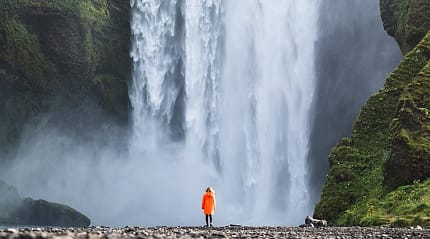 Woman in orange jacket standing at the base of Skogafoss Waterfall in Iceland