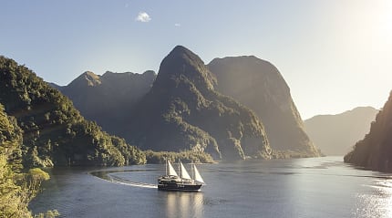 Sailing vessel cruises through tranquil fjord beneath towering forested peaks.