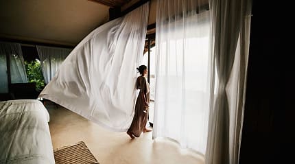 A woman in a flowing dress stands by large windows as a sheer white curtain billows in a luxury hotel room.