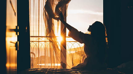 Woman sitting on a bed and opening curtains as the morning sunlight streams into a hotel room.