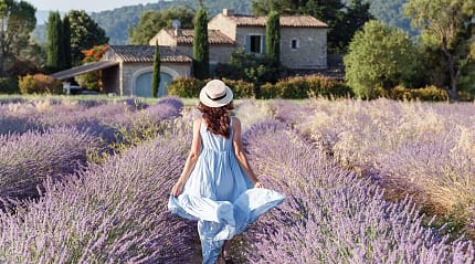 Woman in blue dress walking through a lavender field in Luberon, France, with rustic stone house behind