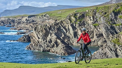 Senior cyclist biking along scenic Irish coastal cliffs with blue ocean waves and rugged landscapes