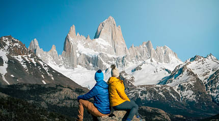 wo hikers in colorful jackets admire snowy Mount Fitz Roy in Patagonia under a clear blue sky
