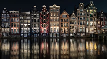 Illuminated traditional Dutch gabled houses reflecting in an Amsterdam canal at night.