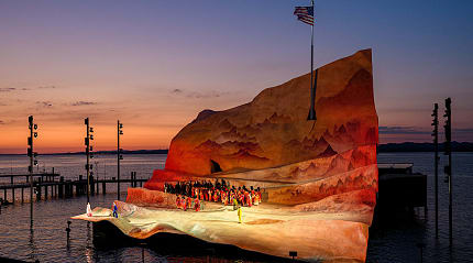 Open-air opera performance on a floating stage at sunset with a dramatic backdrop and calm waters