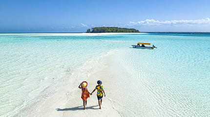 Couple walks on sandbank in clear turquoise water toward island in Zanzibar, Tanzania.