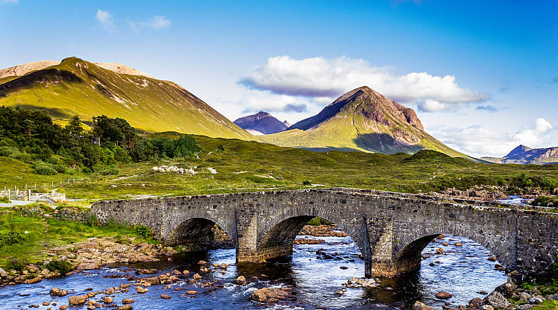 Old vintage brick bridge crossing river in Sligachan, Isle of Skye, Scotland with blue sky, hills and mountains in the background