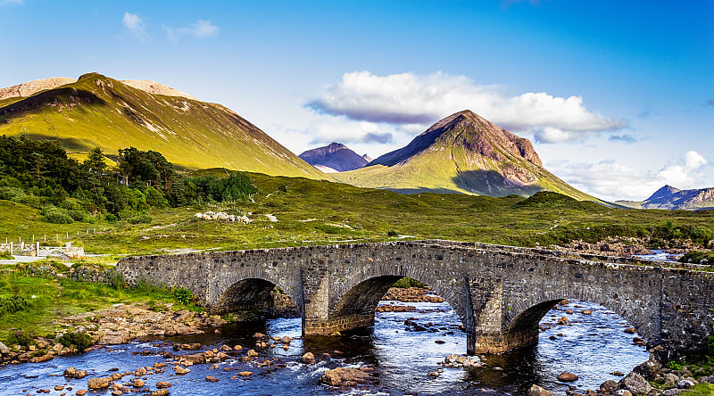 Old vintage brick bridge crossing river in Sligachan, Isle of Skye, Scotland with blue sky, hills and mountains in the background