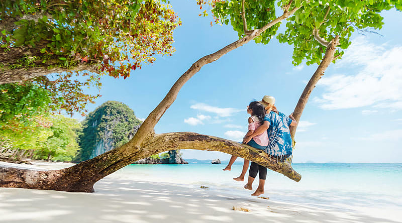 Mother and daughter sitting on a low tree branch on a beach in Phuket, Thailand