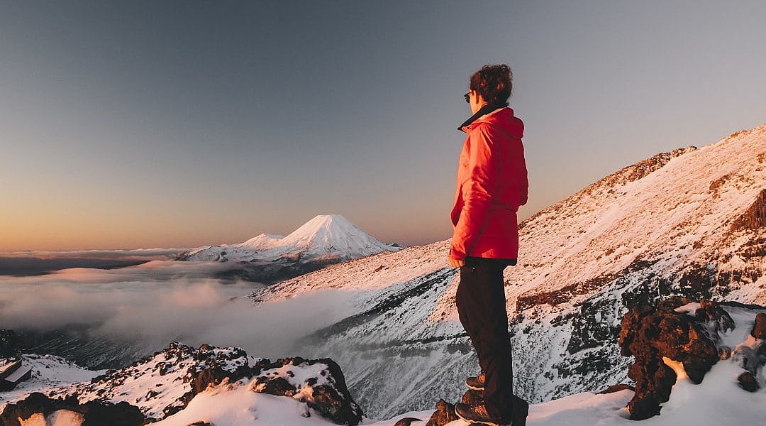 Hiker in winter jacket standing on snowy mountain, admiring view of snow-capped peaks and clouds at sunrise in Tongariro National Park, New Zealand