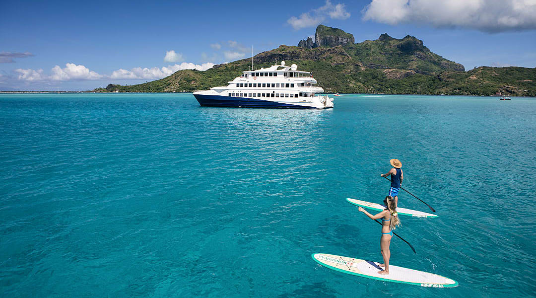 Couple paddleboarding on turquoise water in Bora Bora with a luxury yacht and tropical island backdrop