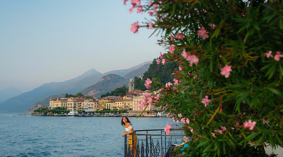 Scenic view of a woman by Lake Como, vibrant flowers, waterfront architecture, and mountains in the distance