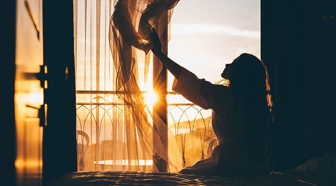 Woman sitting on a bed and opening curtains as the morning sunlight streams into a hotel room.