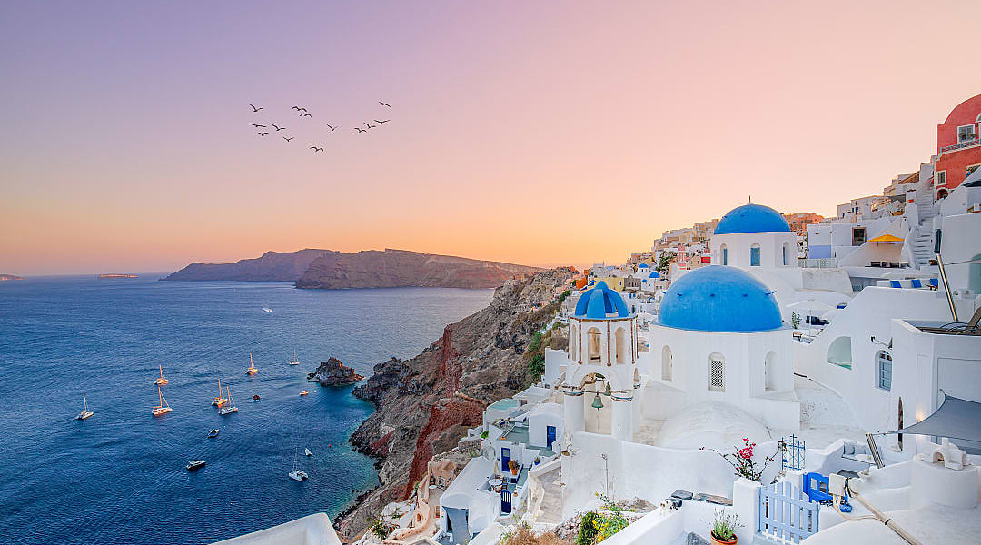 The sun sets over Oia’s blue-domed churches and Aegean Sea.