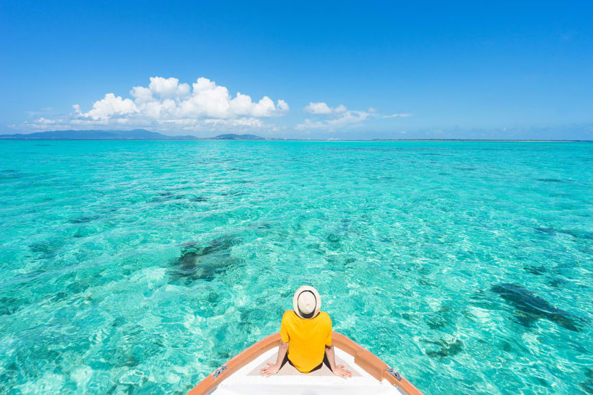 Man sitting on the front of a boat sailing through a clear turquoise water on a perfect summer vacation day around tropical islands of Okinawa, japan