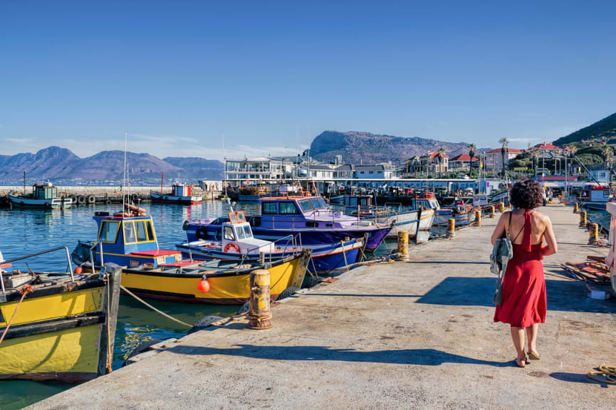 Woman walking on pier with colorful fishing boats in Kalk Bay, South Africa