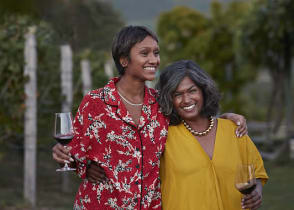 Smiling mother and daughter holding wineglasses and standing with arms around each other at a vineyard