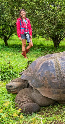 Smiling woman standing near giant tortoise in lush greenery.