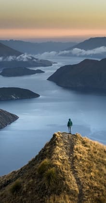 Person standing atop mountain ridge overlooking lakes and distant hills.