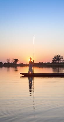 Man standing on canoe at sunrise over calm reflective water.