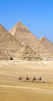 Camels and riders crossing desert with pyramids in the background.