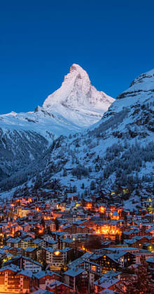 Snowy mountain towers above illuminated alpine village at twilight.