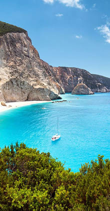 Sailboat floats near steep cliffs and turquoise beach in sunlight.