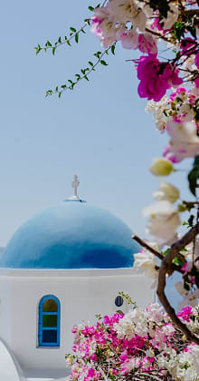 A traditional white Greek building with a vibrant blue dome and a cross on top, framed by bright pink and white bougainvillea flowers, set against a clear blue sky