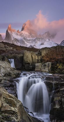 A cascading waterfall and rocky stream in the foreground, with the snow-capped peaks of Mount Fitz Roy illuminated by pink light in the background, in Los Glaciares National Park, Argentina.