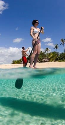 Couple paddleboarding in clear turquoise water near tropical beach.