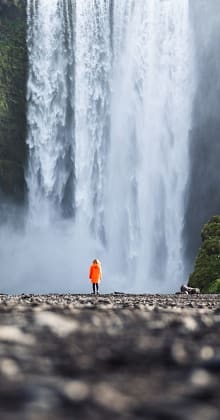 Hiker in orange coat stands before massive cascading waterfall spray.