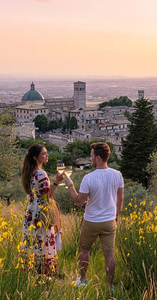 Couple toasting with wine in a field, overlooking Assisi, Italy, at sunset.